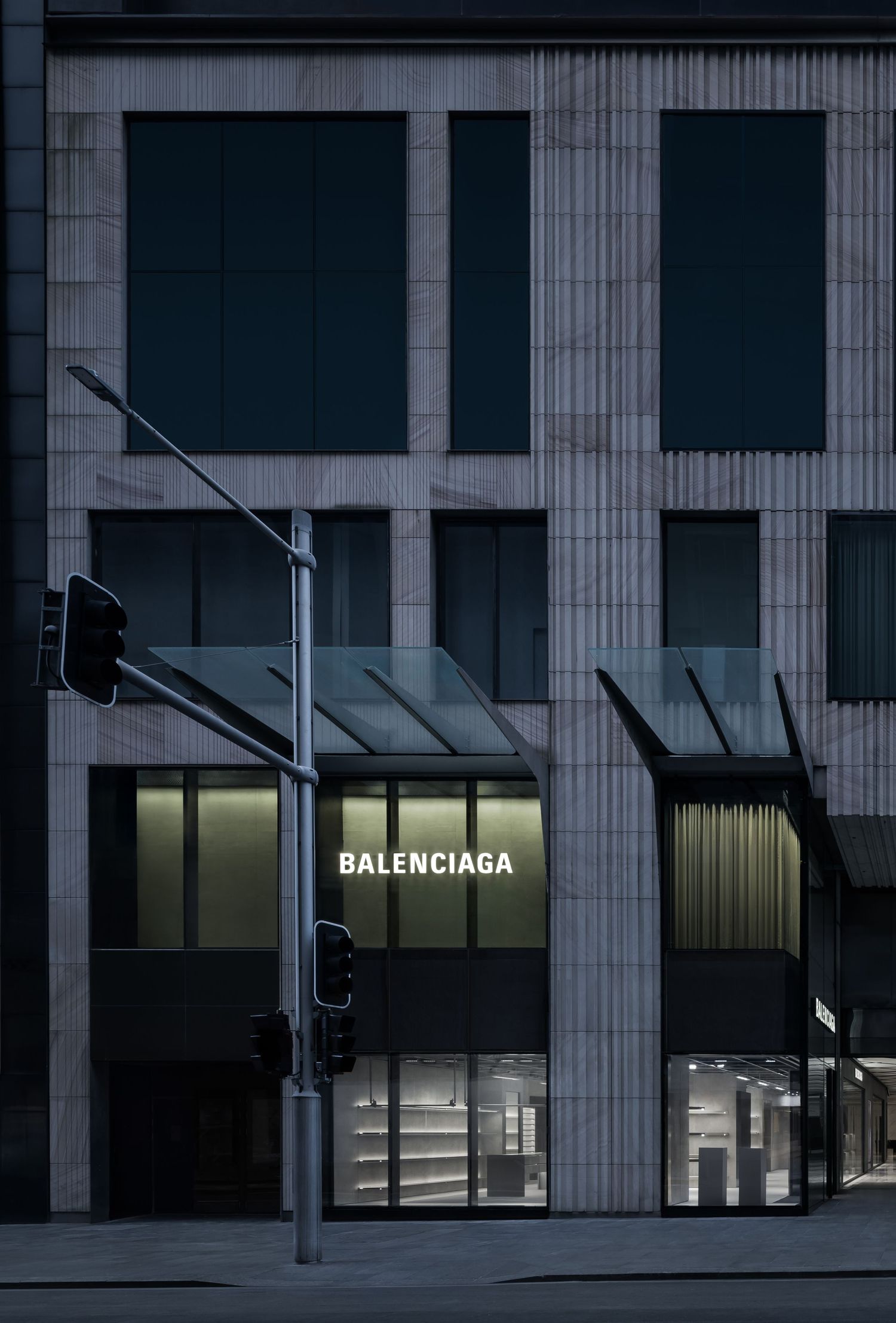 Balenciaga flagship storefront on a city street corner, featuring a stone facade, glass canopies, and softly lit window displays at dusk.