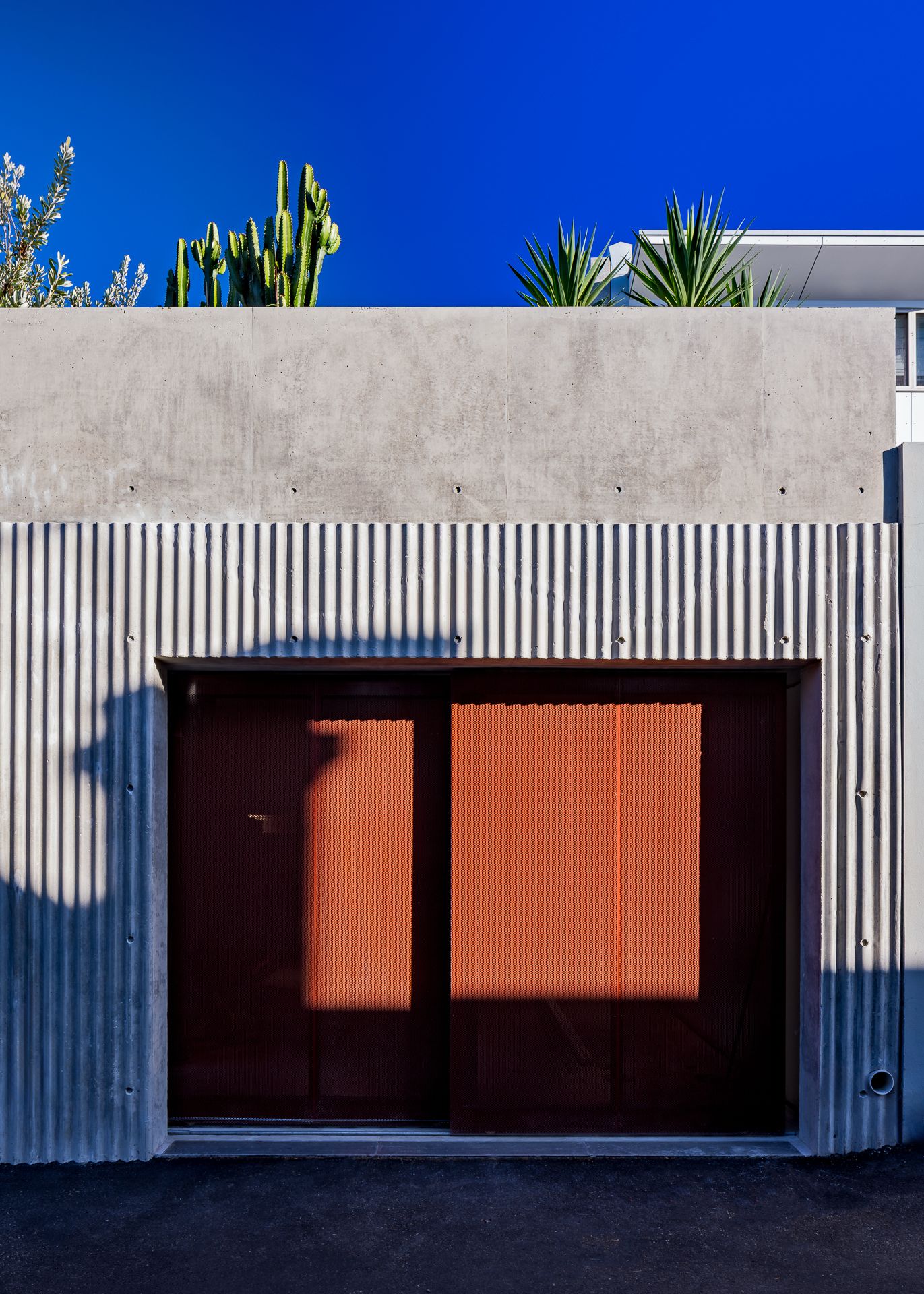 Concrete and corrugated metal facade with a dark garage door and rust-colored panels beneath a vivid blue sky and cacti.