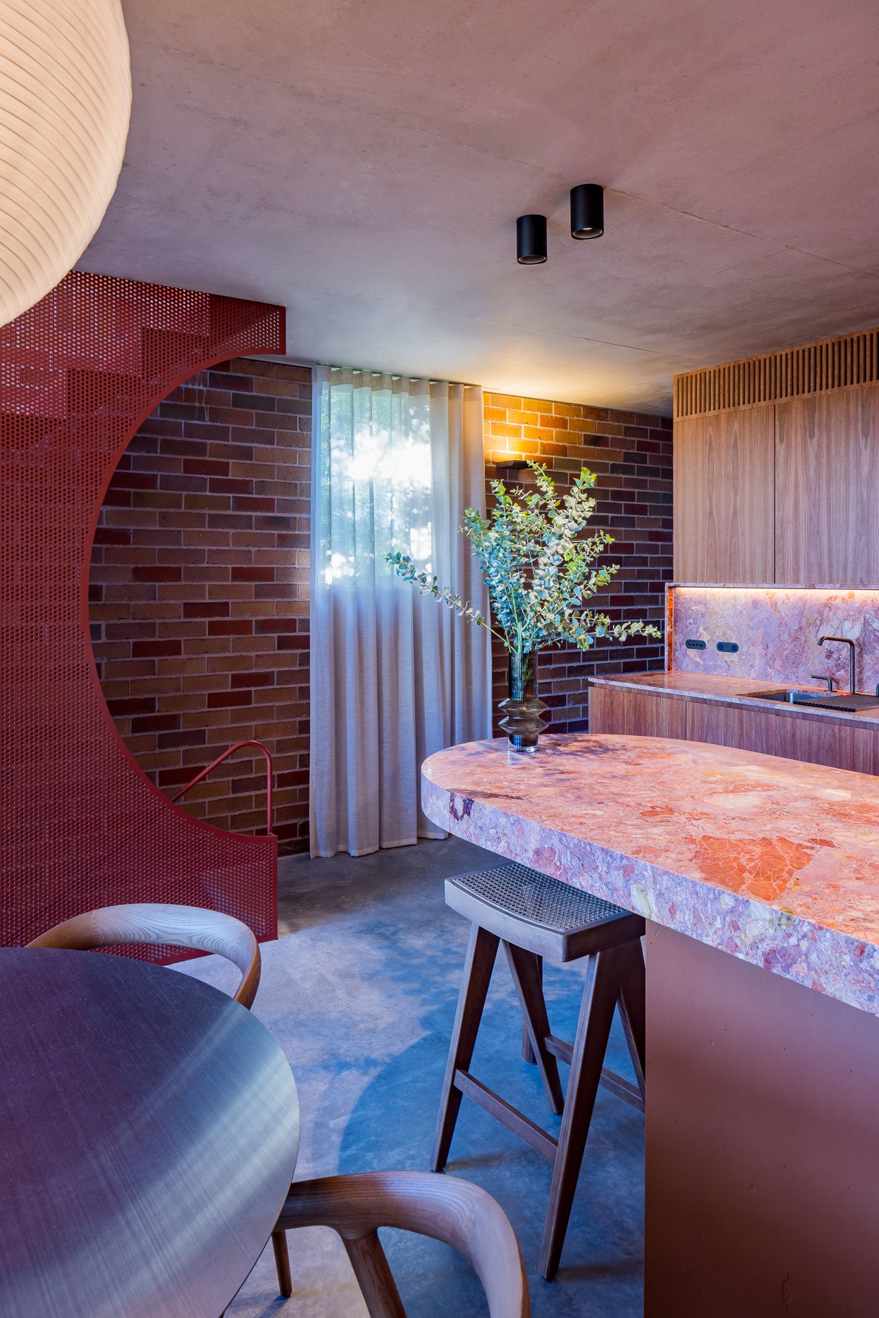 Modern open-plan kitchen with pink marble island, timber stools, red brick walls and a curved red perforated metal screen.