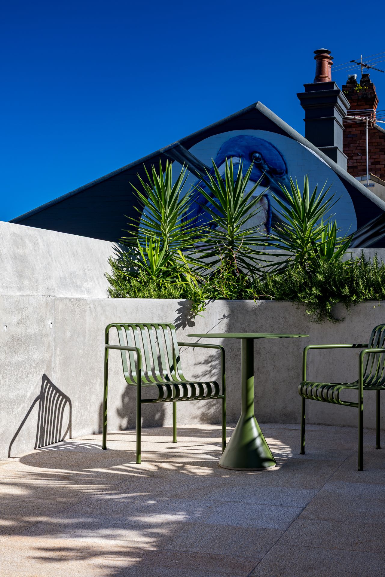 Green metal outdoor chairs and a round table on a sunlit concrete terrace with spiky plants and a mural-covered roofline against a blue sky.