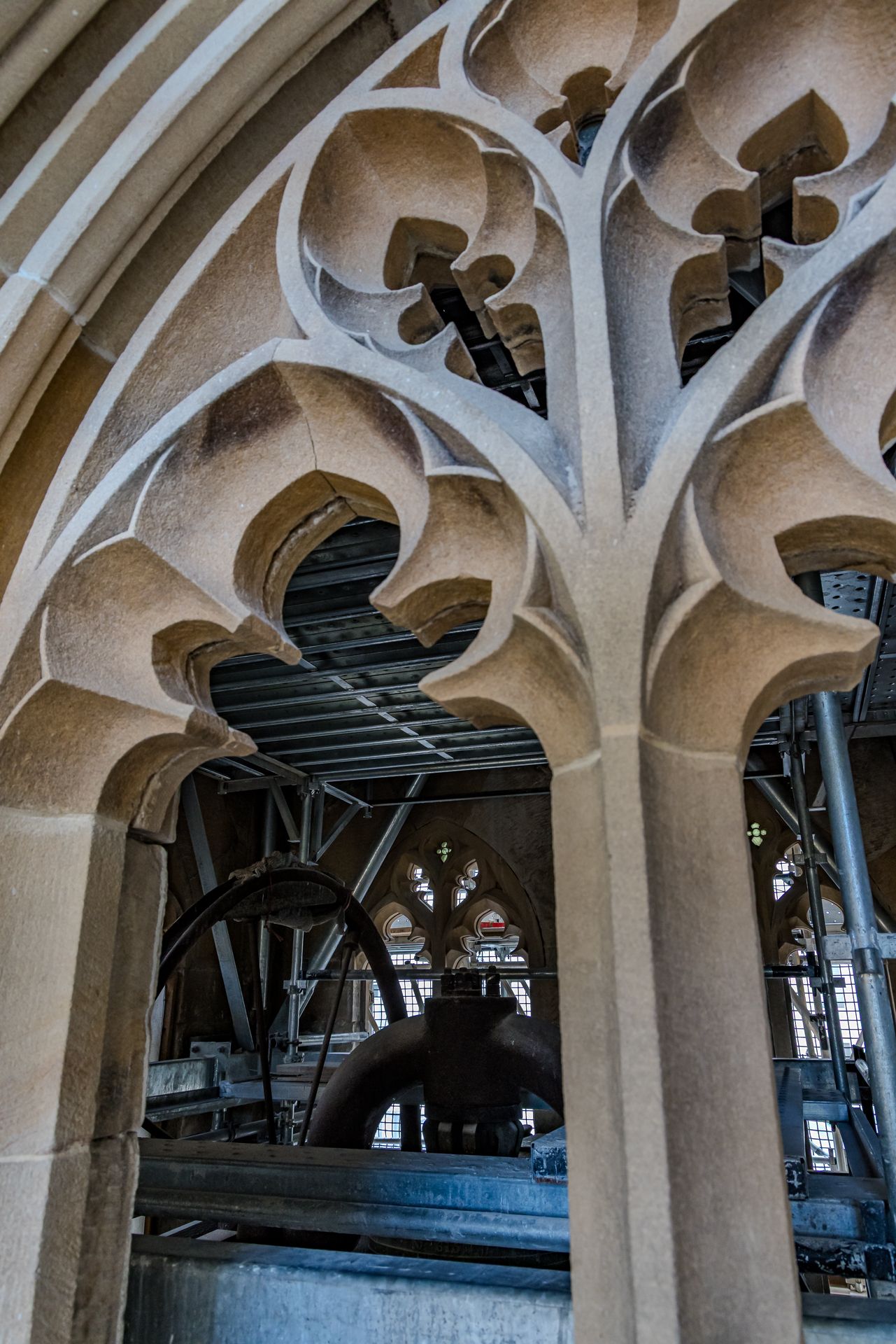 Gothic stone tracery with cusped arches framing a view of bell tower machinery and steel supports.