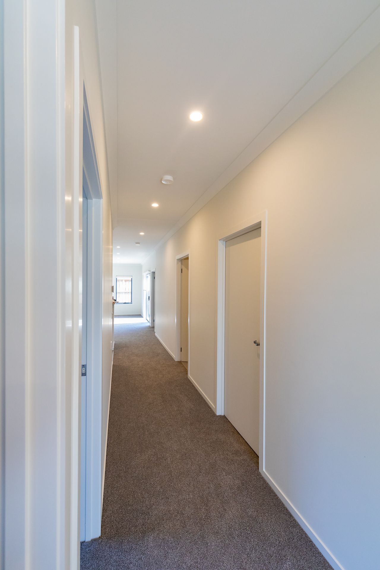 Long hallway with grey carpet, white walls, recessed ceiling lights and several closed doors leading to rooms in a new home.