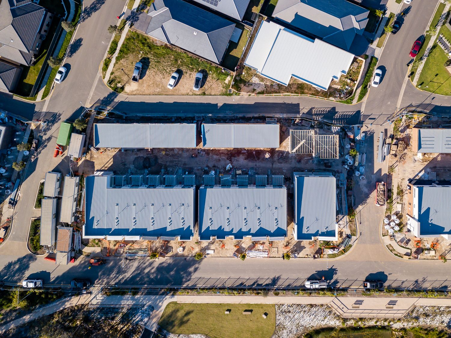 Aerial top-down view of newly built townhouses with blue metal roofs surrounded by streets, parked cars and neighbouring suburban homes.