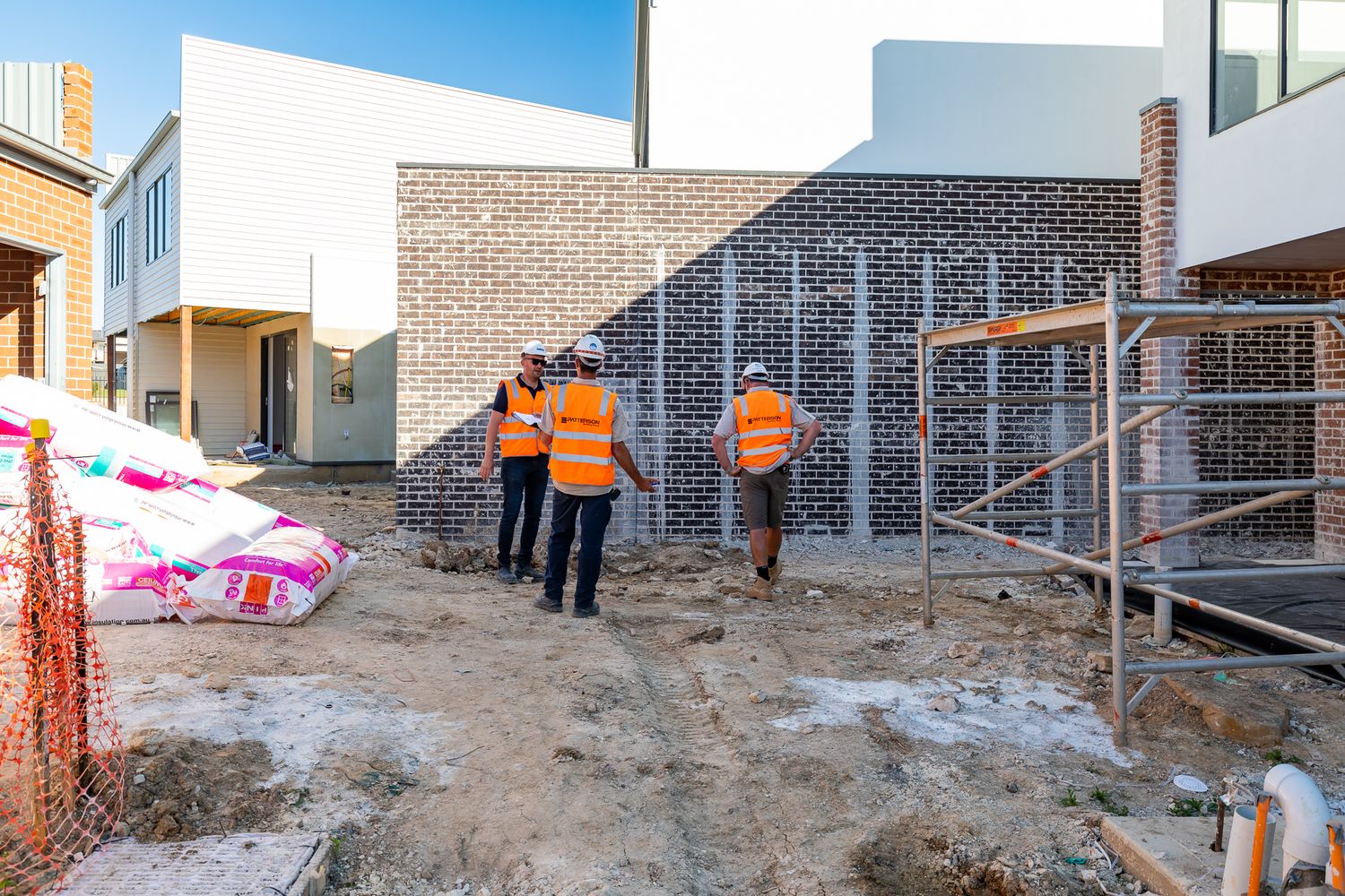 Three workers in orange hi-vis vests walk across a muddy site toward a brick wall near scaffolding and new buildings.