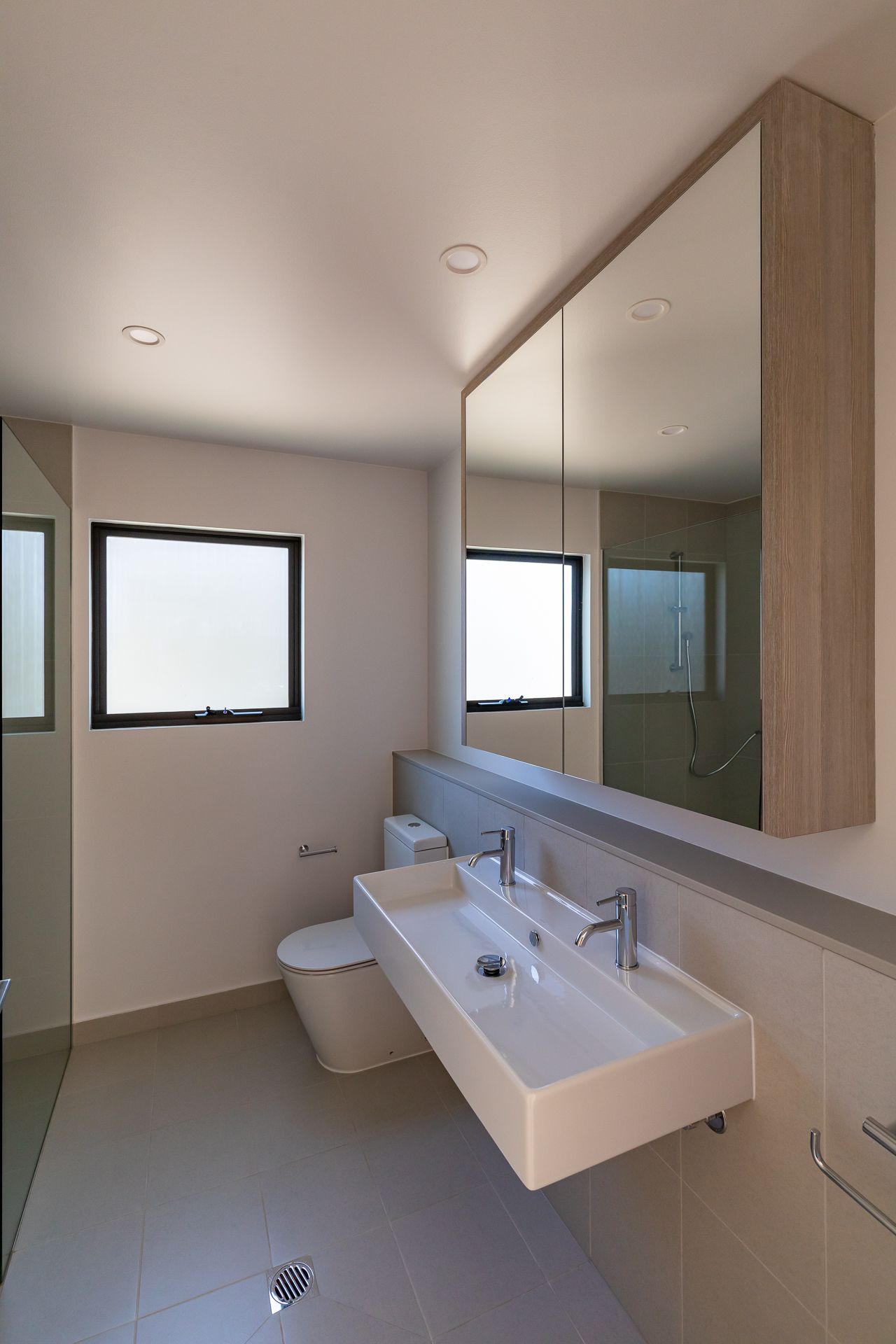 Modern bathroom with a wall-mounted white basin, mirrored cabinet, frosted window, toilet and tiled floor in a newly built home.