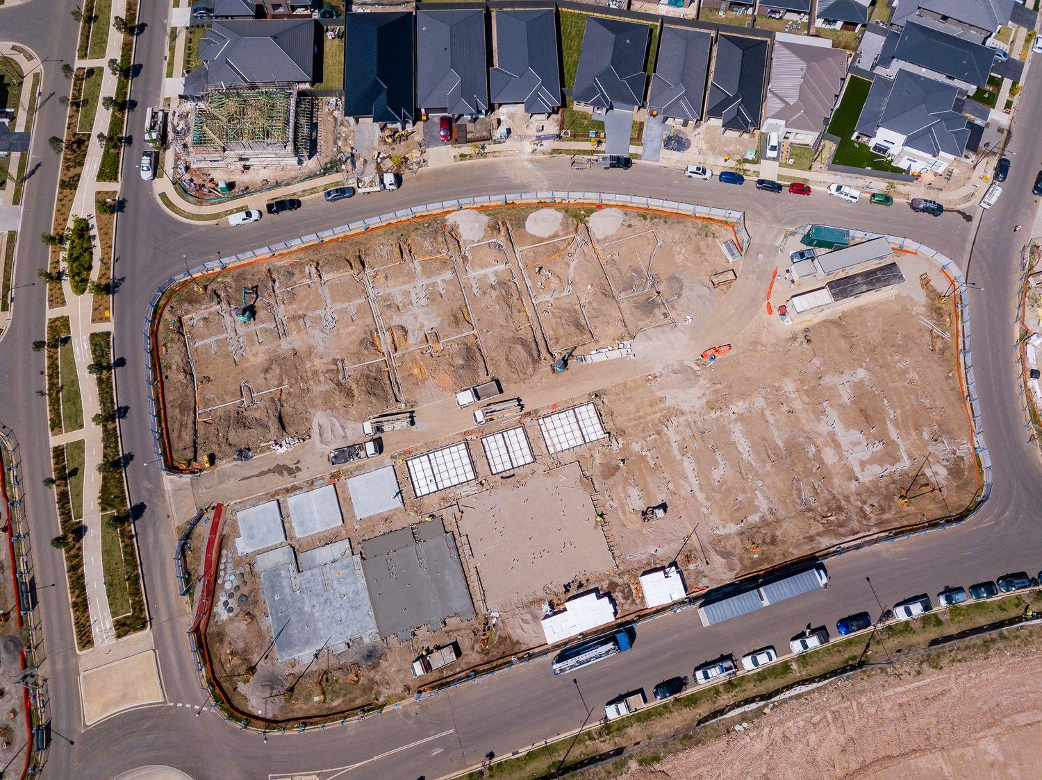 Aerial view of a residential construction site with excavated lots, building materials, and parked vehicles surrounded by roads and new home