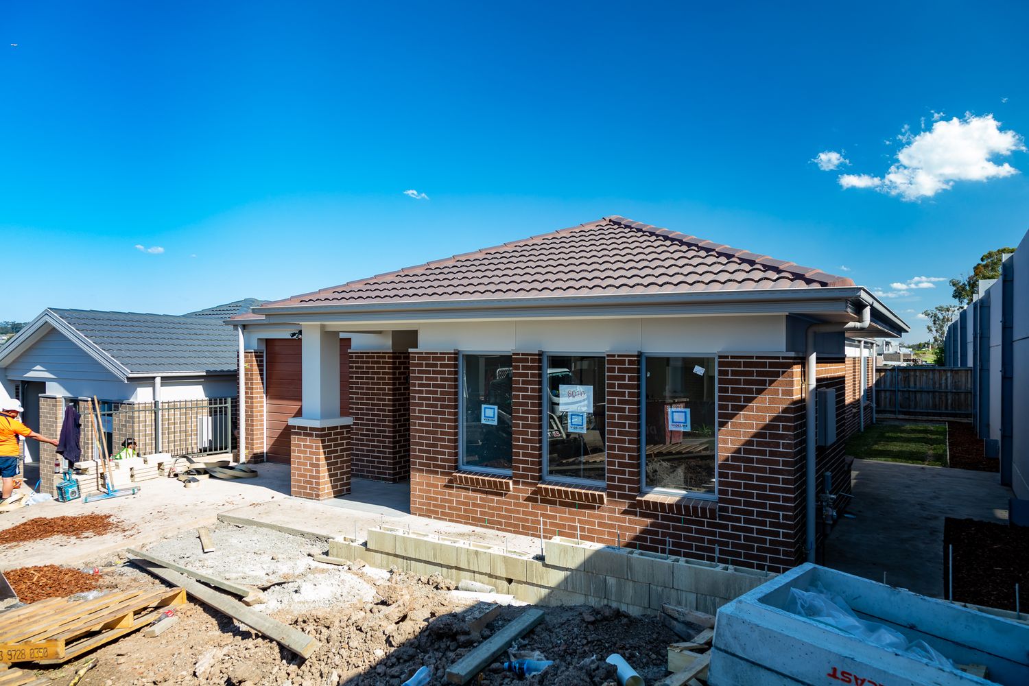 Newly built brick home with a tiled roof under construction, with building materials and rubble in the foreground beneath a blue sky.