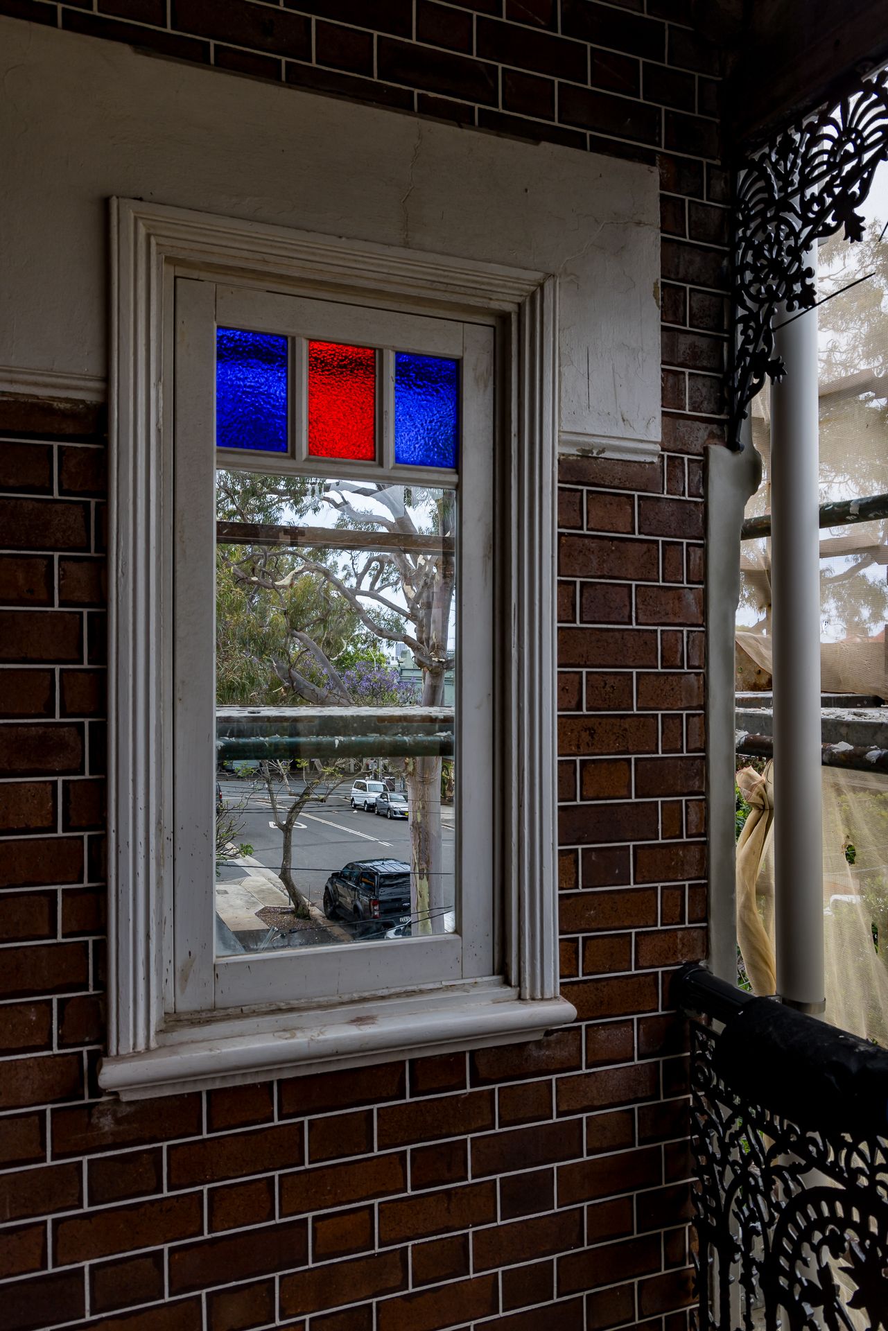 Heritage brick wall with a timber-framed window featuring red and blue stained glass panes beside a cast iron lace balcony.