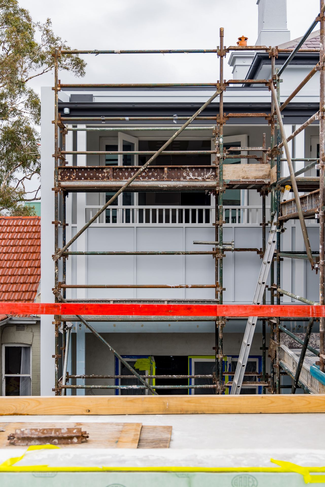 Home renovation in progress with scaffolding around a grey two-storey house, a covered balcony and timber framing in the foreground.