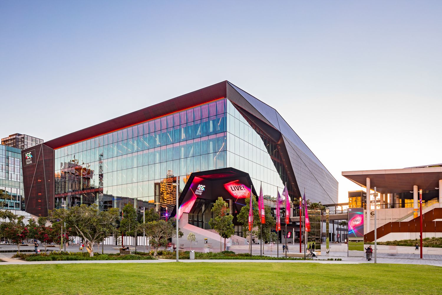 Large contemporary glass and steel convention building with a sharp angular roof beside a green lawn at sunset.