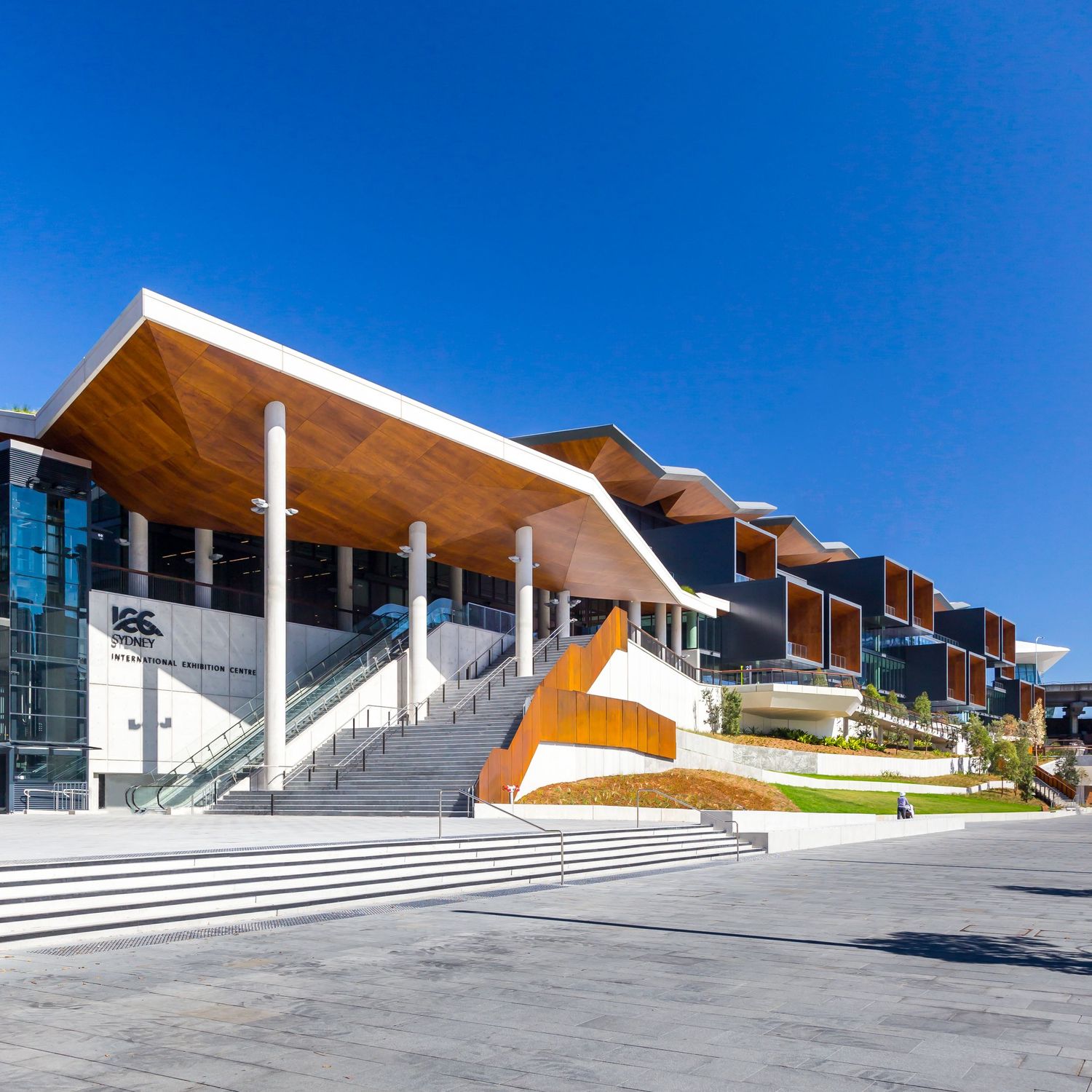 Contemporary building entrance with a timber-clad canopy, wide stone steps and white columns beneath a bright blue sky.