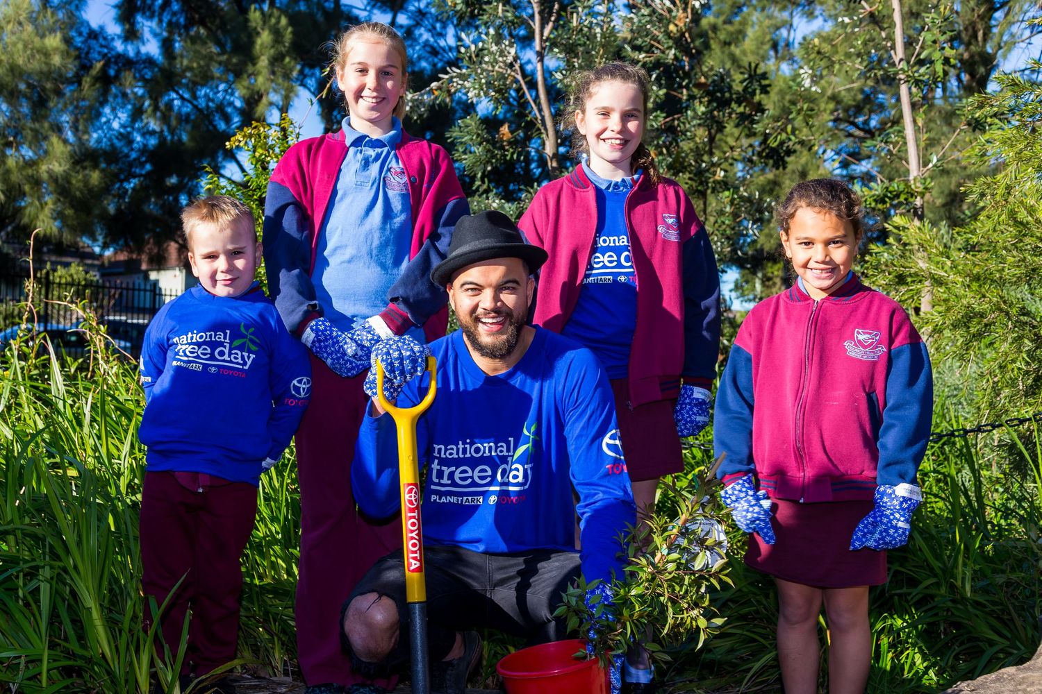 A man kneels with a shovel among four children in National Tree Day shirts, all wearing gloves in a green planting area.