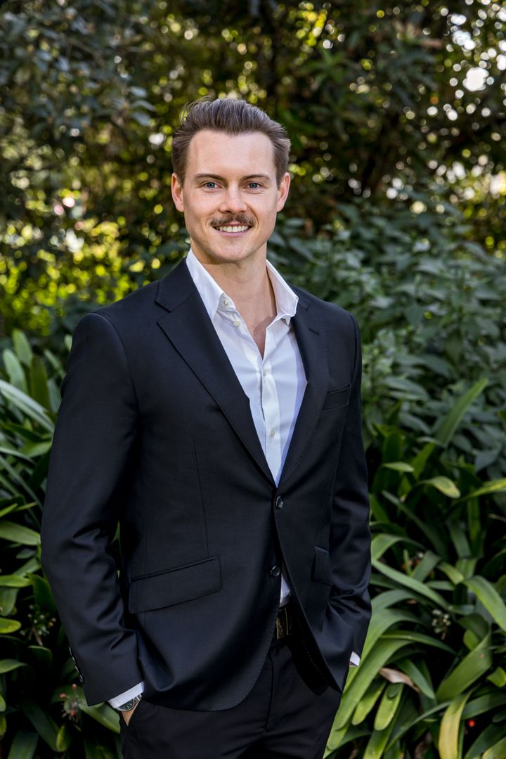 Smiling man with a mustache wearing a navy blazer and white shirt standing outdoors in front of lush green foliage.
