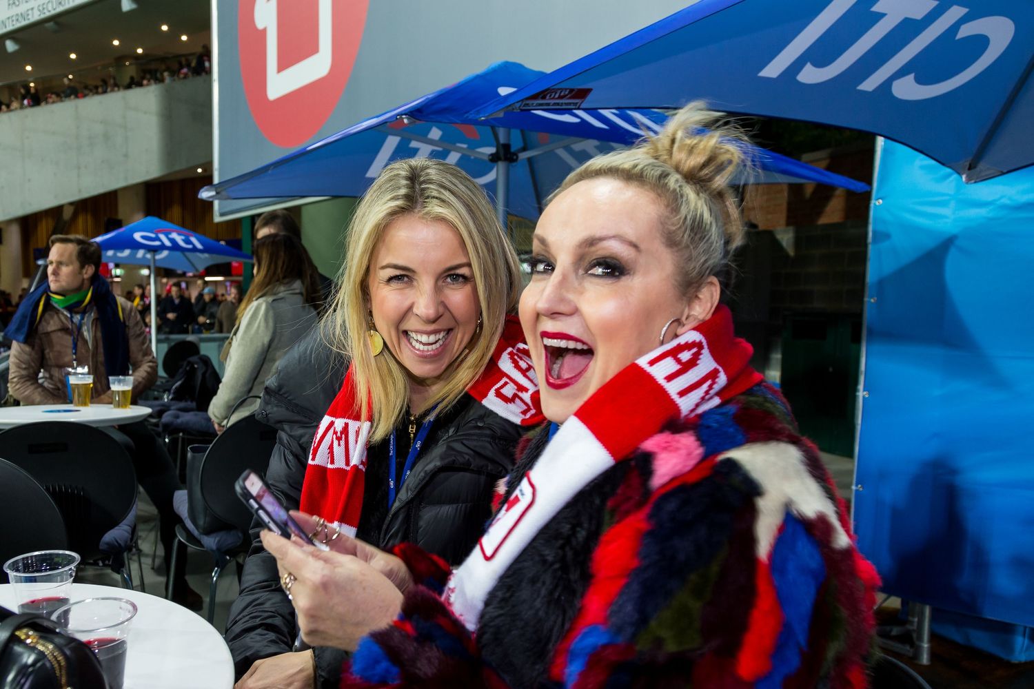 Two cheerful women in red and white scarves pose for a photo at an outdoor event with umbrellas and tables behind them.