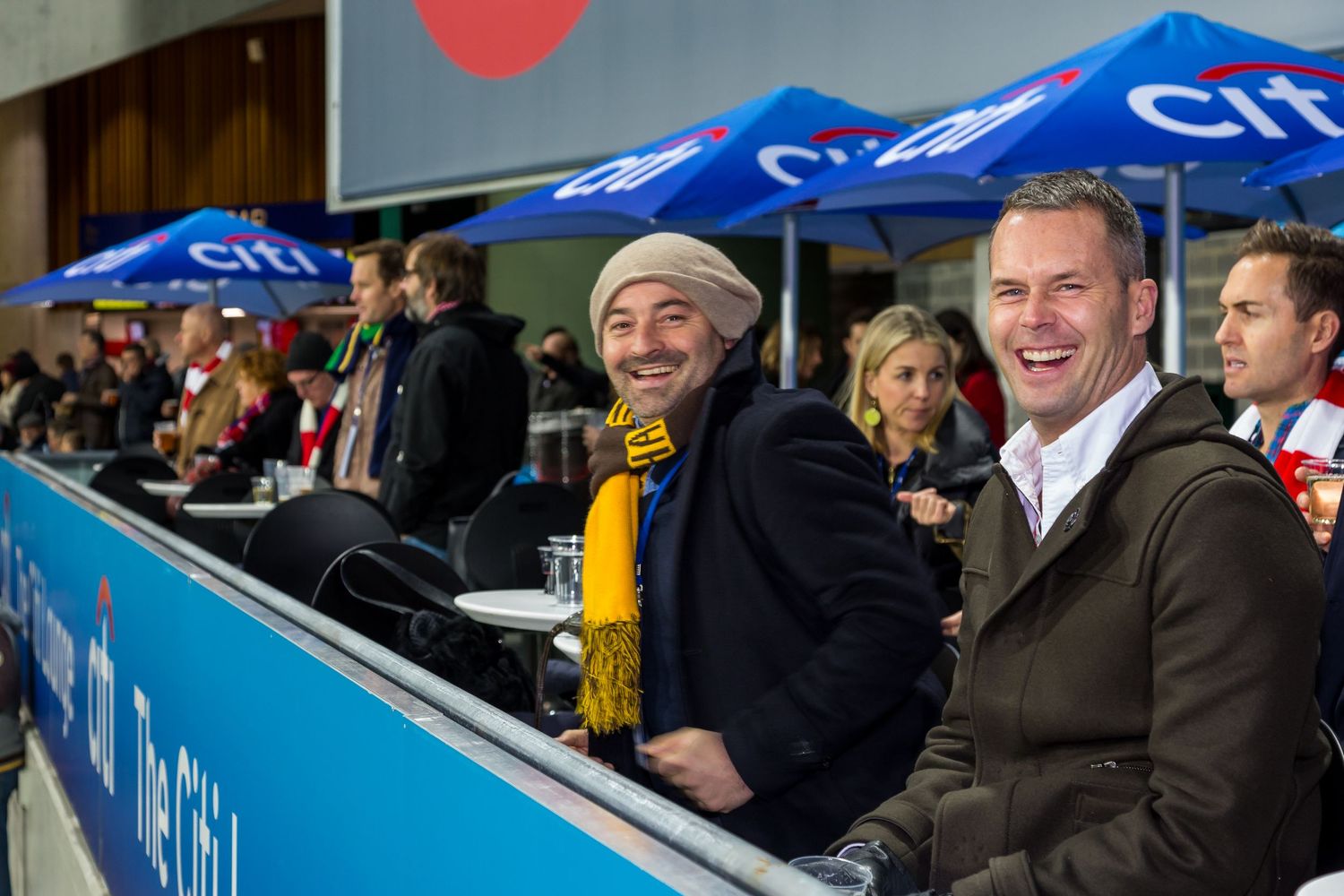 Two smiling men in jackets sit in a stadium seating area watching an event, with blue umbrellas and spectators behind them.