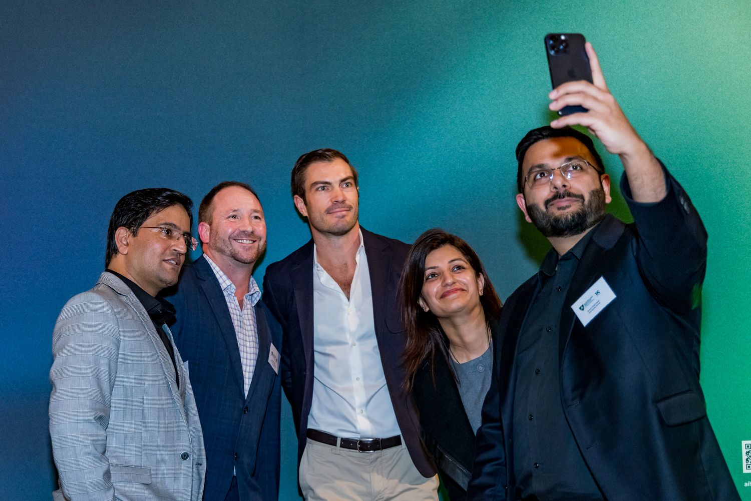 Four smiling people pose close together for a group selfie while a bearded man in glasses holds up a smartphone at an event.