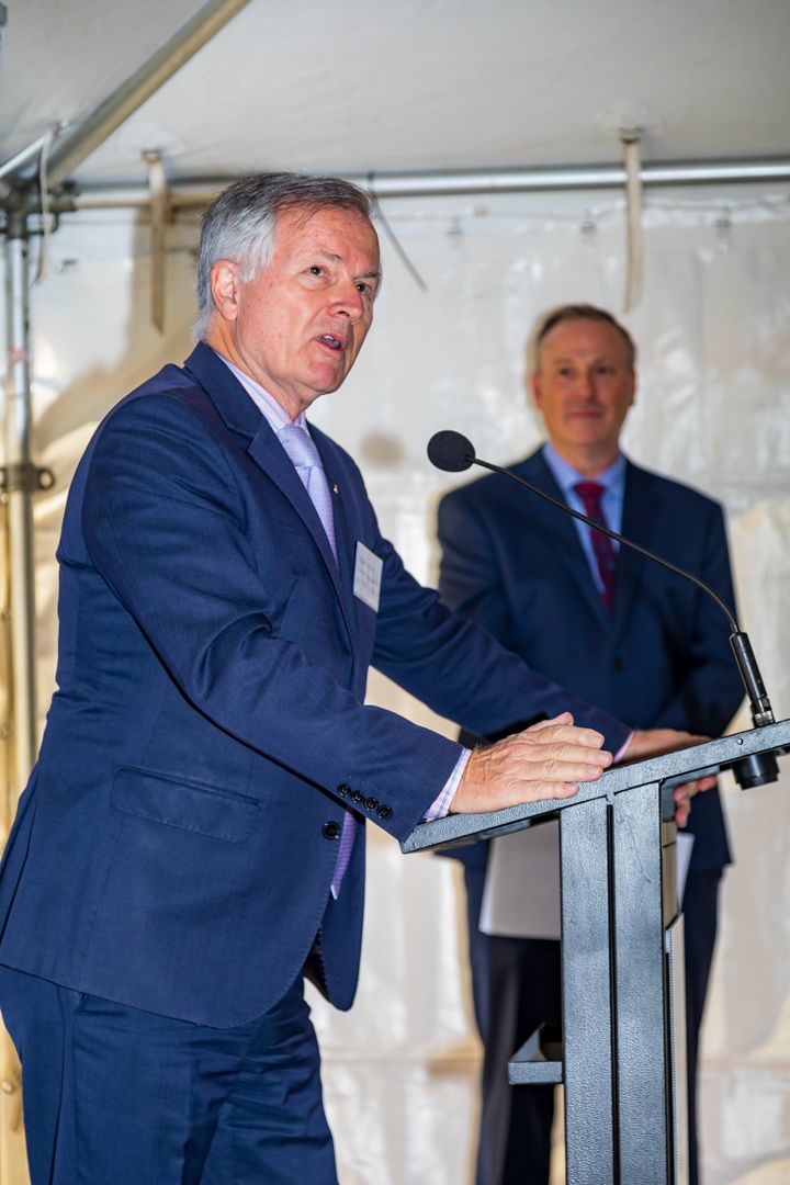 A man in a navy blazer speaks at a podium with a microphone while another man in a suit watches in the background.