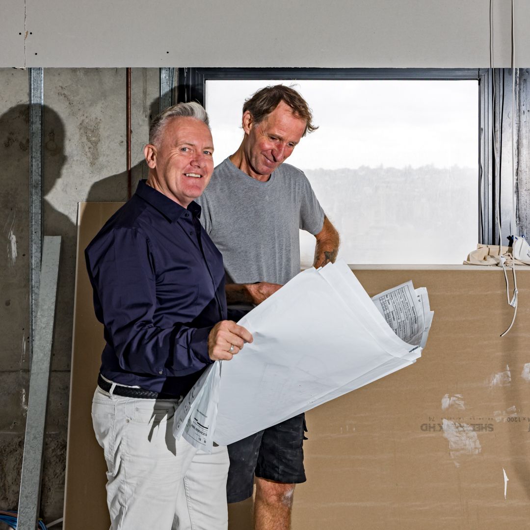 Two men smiling and reviewing printed building plans inside an unfinished construction space with a window view behind them.