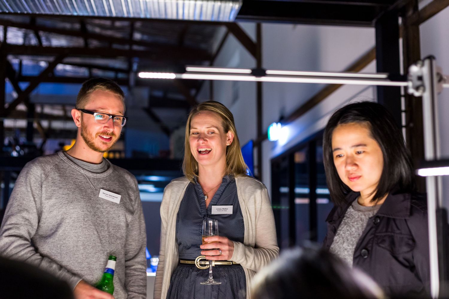 Three people chat and smile at an evening networking event inside an industrial-style venue, holding drinks and wearing name tags.