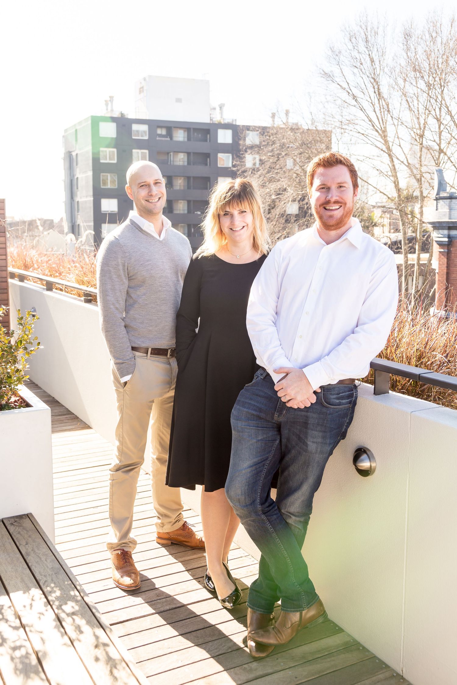 Two men and a woman smile together on a sunlit rooftop terrace with planters and city buildings in the background.