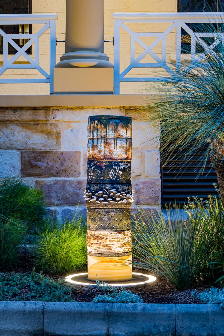 Illuminated stacked ceramic sculpture with a glowing ring base in a landscaped garden beside a sandstone wall at dusk.
