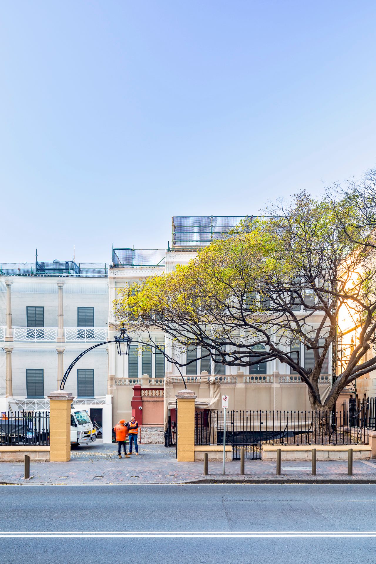 Two workers in orange high-visibility vests stand beside a truck near a historic building under renovation, with a leafy tree nearby.