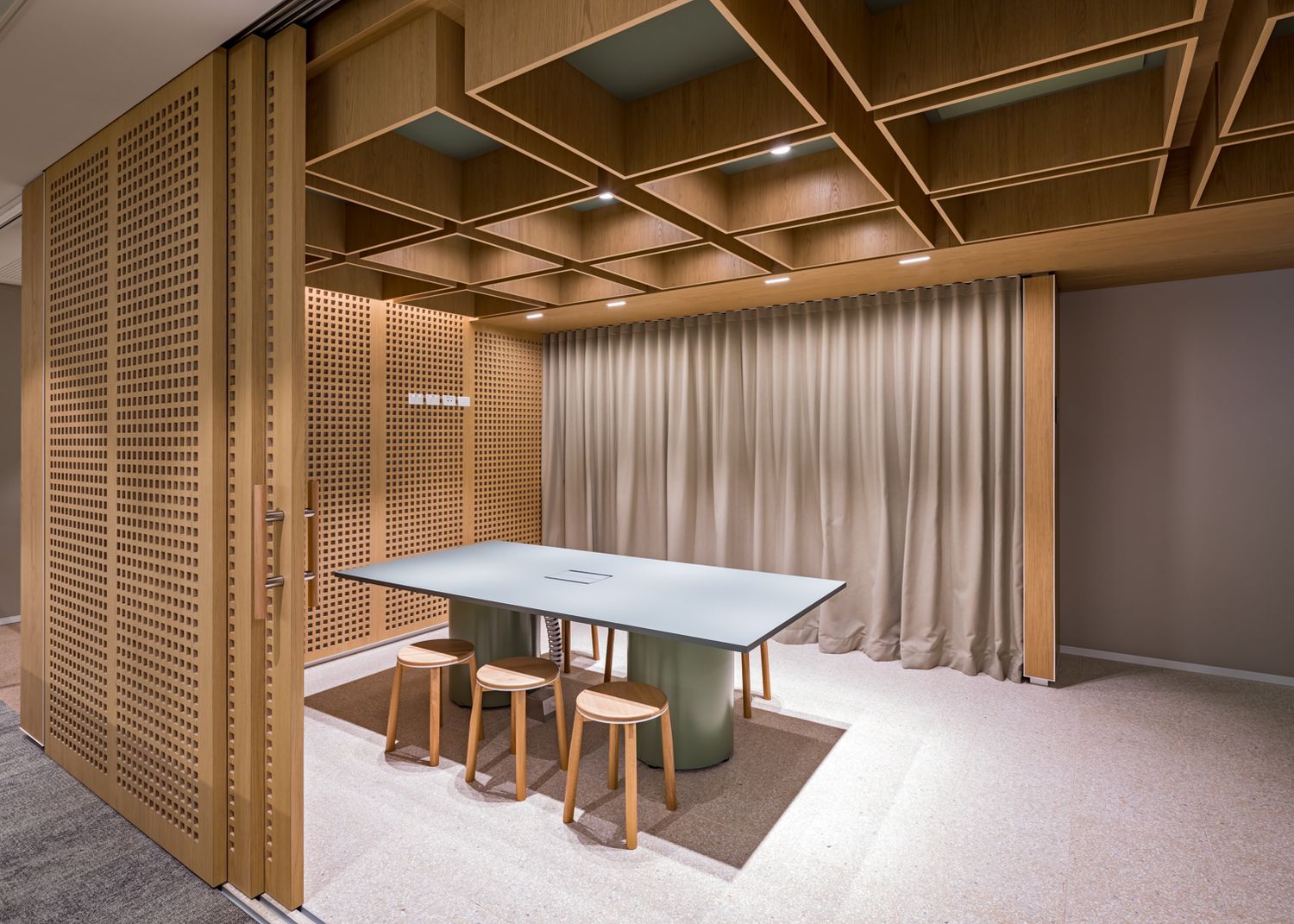 Contemporary meeting room featuring a wooden slatted door, grid timber ceiling, taupe curtains and a green-topped table with stools.