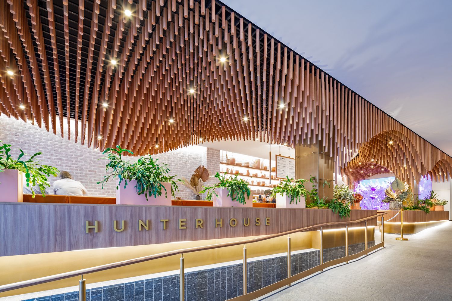 Hunter House cafe entrance with a curved counter beneath a flowing timber tube ceiling, lush plants, and a brick feature wall.