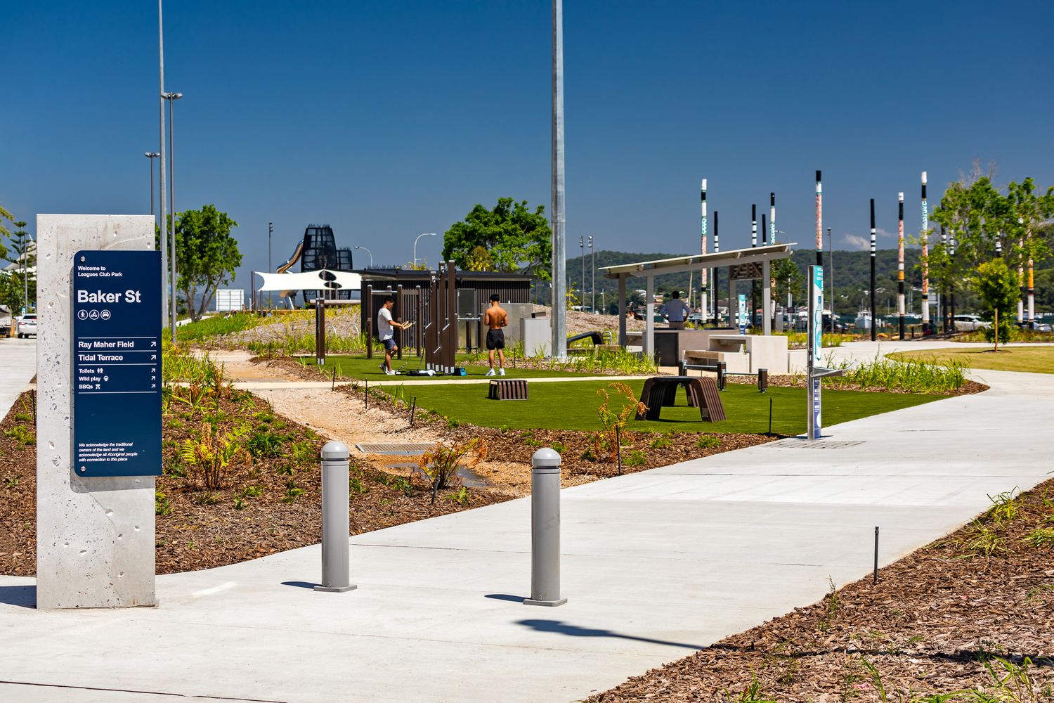 Park signpost beside a curved concrete path with bollards, painted poles, lawn and a play area under a clear blue sky.