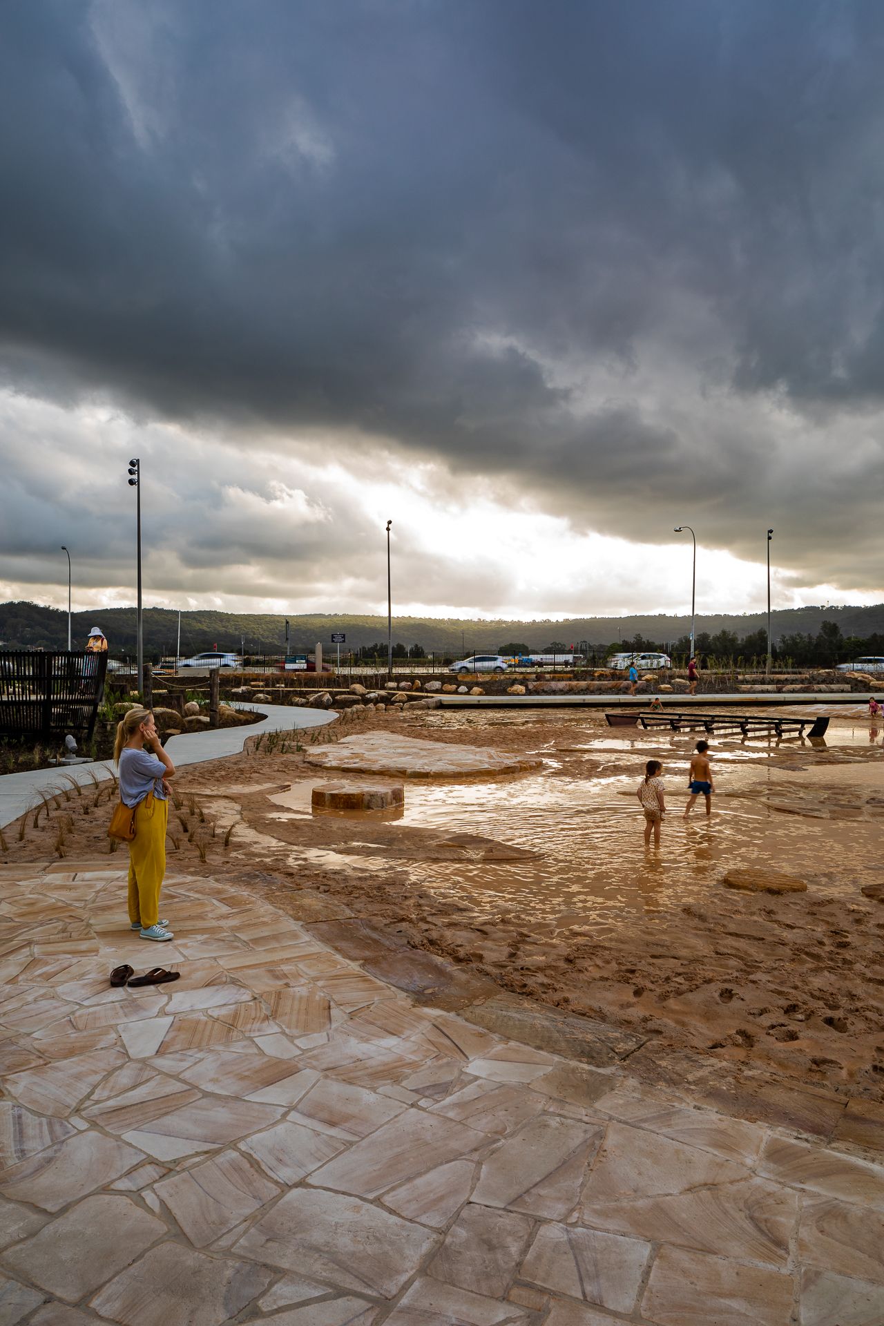 Woman in yellow trousers photographs a waterfront park under dramatic storm clouds as children play in shallow water on sandstone paving.