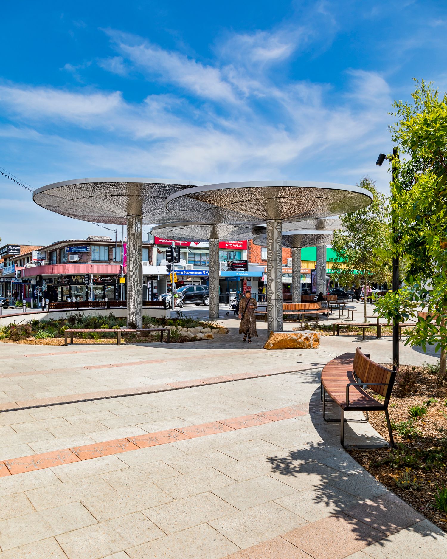 Modern public plaza with large perforated metal canopy shades, wooden benches, paved walkways and shopfronts under a bright blue sky.
