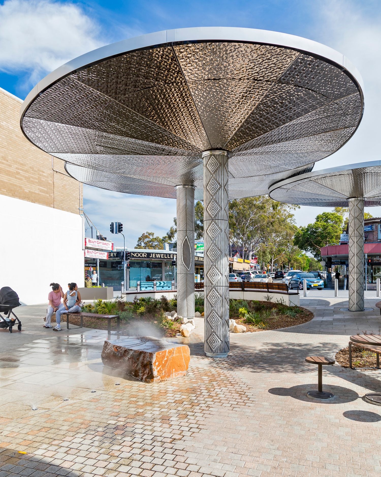 Perforated metal umbrella canopy with a patterned column shades a paved town square, with people seated on a bench near street shops.