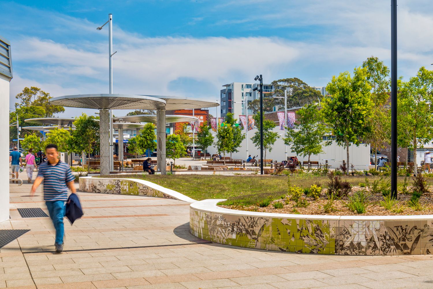 A pedestrian walks through a revitalised town plaza with metal shade canopies, a curved mural-decorated garden wall, and green trees.