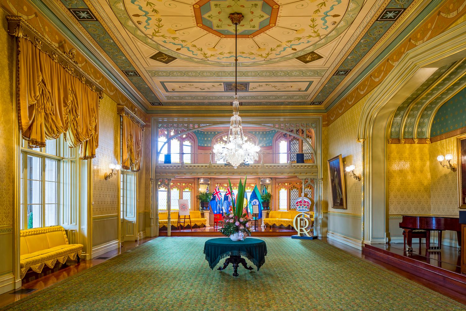 Spacious historic reception hall with a painted ceiling, chandelier, gold curtains, green carpet and a view toward an arched balcony.