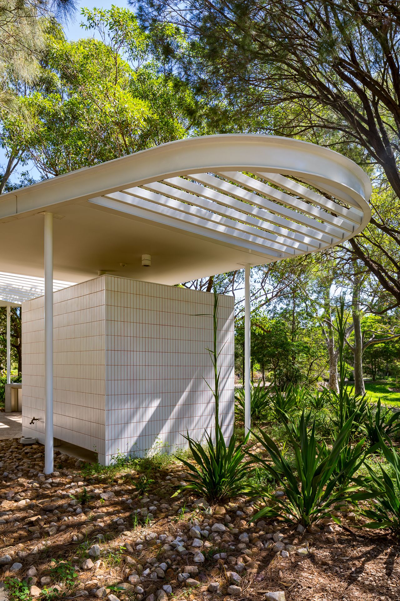 Close view of a white tiled restroom building with a curved slatted roof, agave plants and pebble garden bed.