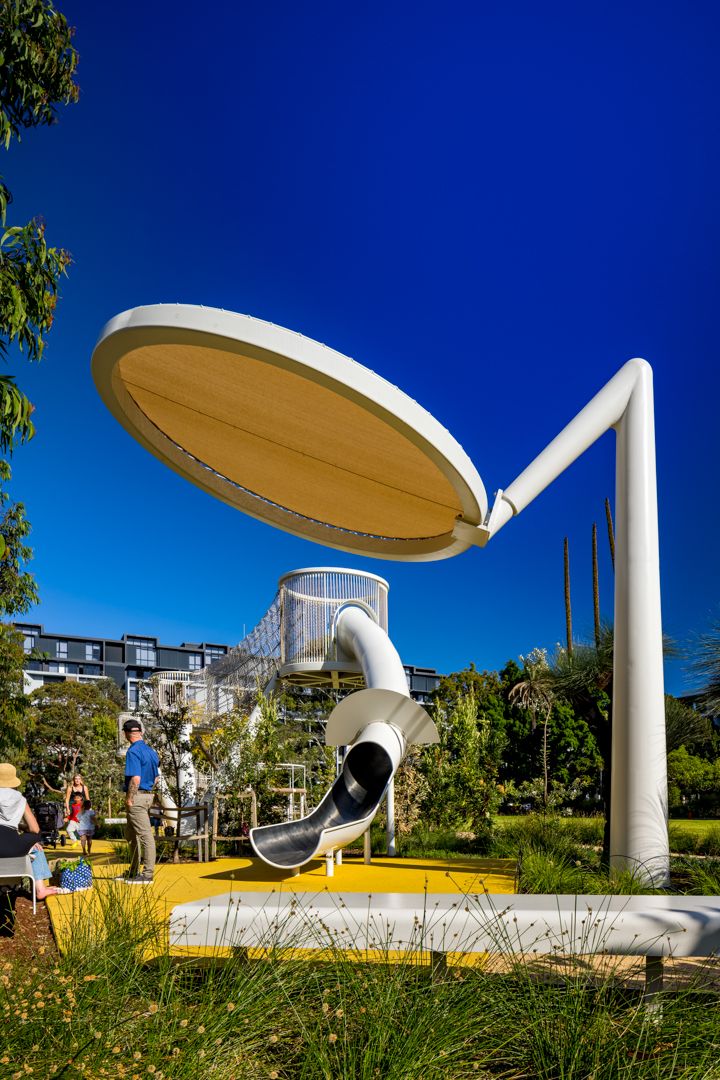Playground with a spiral white slide and a large circular timber-underside canopy on a post, set against a deep blue sky.