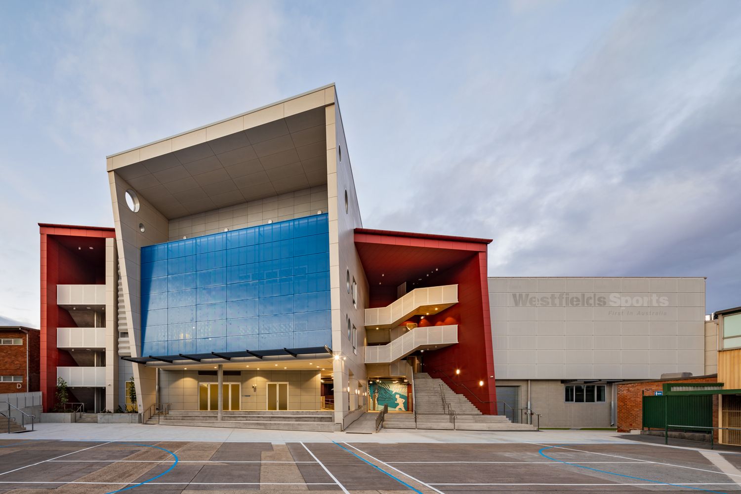Colorful modern school building with red and blue facade, open outdoor staircases, and an angular cantilevered roof over a car park.