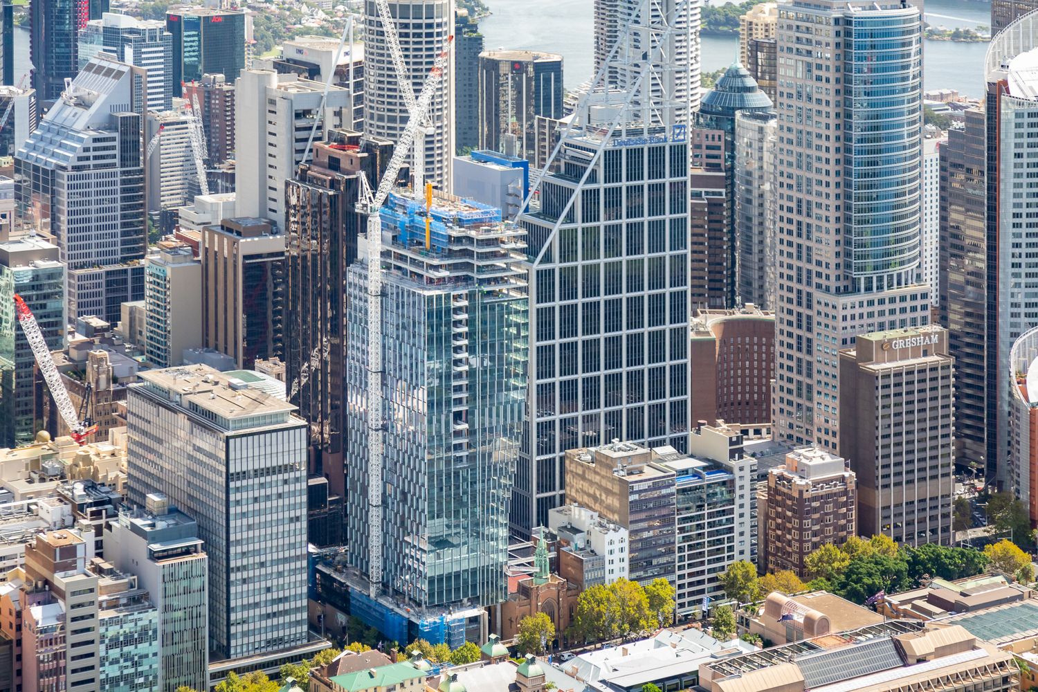 Aerial view of modern glass skyscrapers and mid-rise buildings in a dense city center with a tower crane and harbour glimpse.