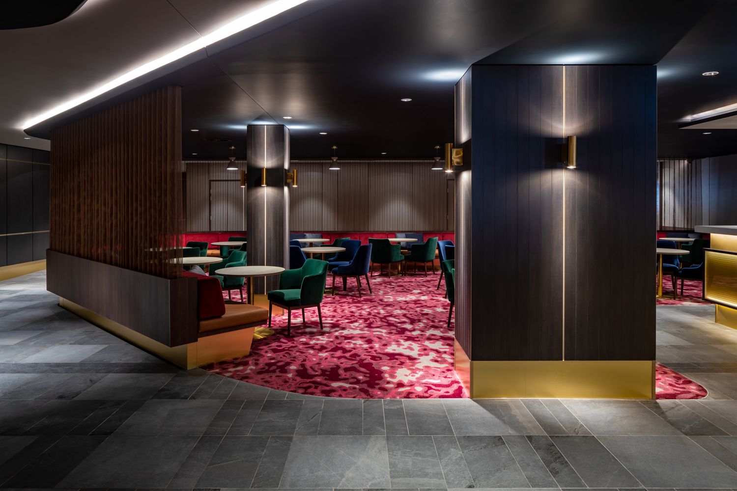 Upscale lounge with dark timber panelling, brass accents, green velvet chairs and a bold pink patterned carpet under moody lighting.