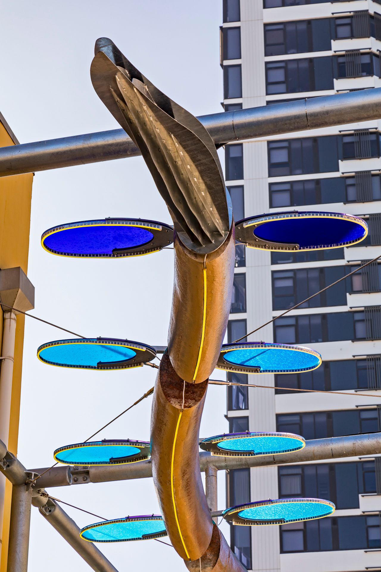 Curved metallic sculpture with blue translucent discs and a glowing yellow strip against a high-rise apartment tower.