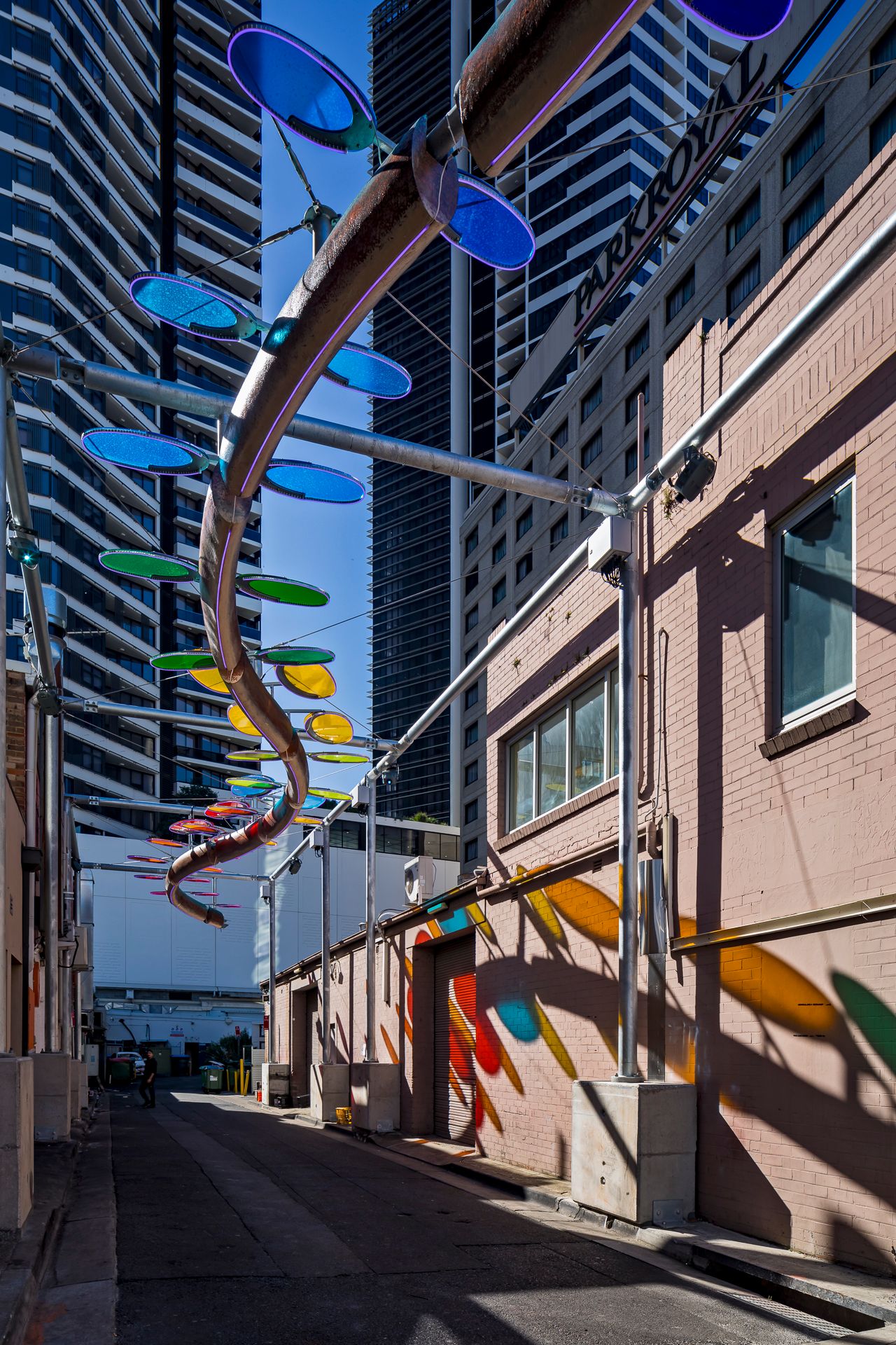 Looking up a narrow laneway at a curving sculpture with blue and green discs, as colorful shadows fall on a brick wall.