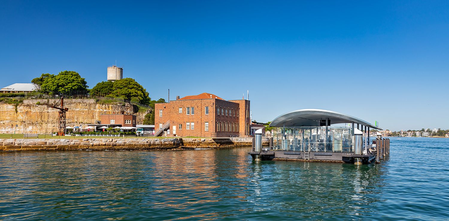 Modern glass-walled ferry terminal on the harbour beside a red brick building and a sandstone wall under a deep blue sky.