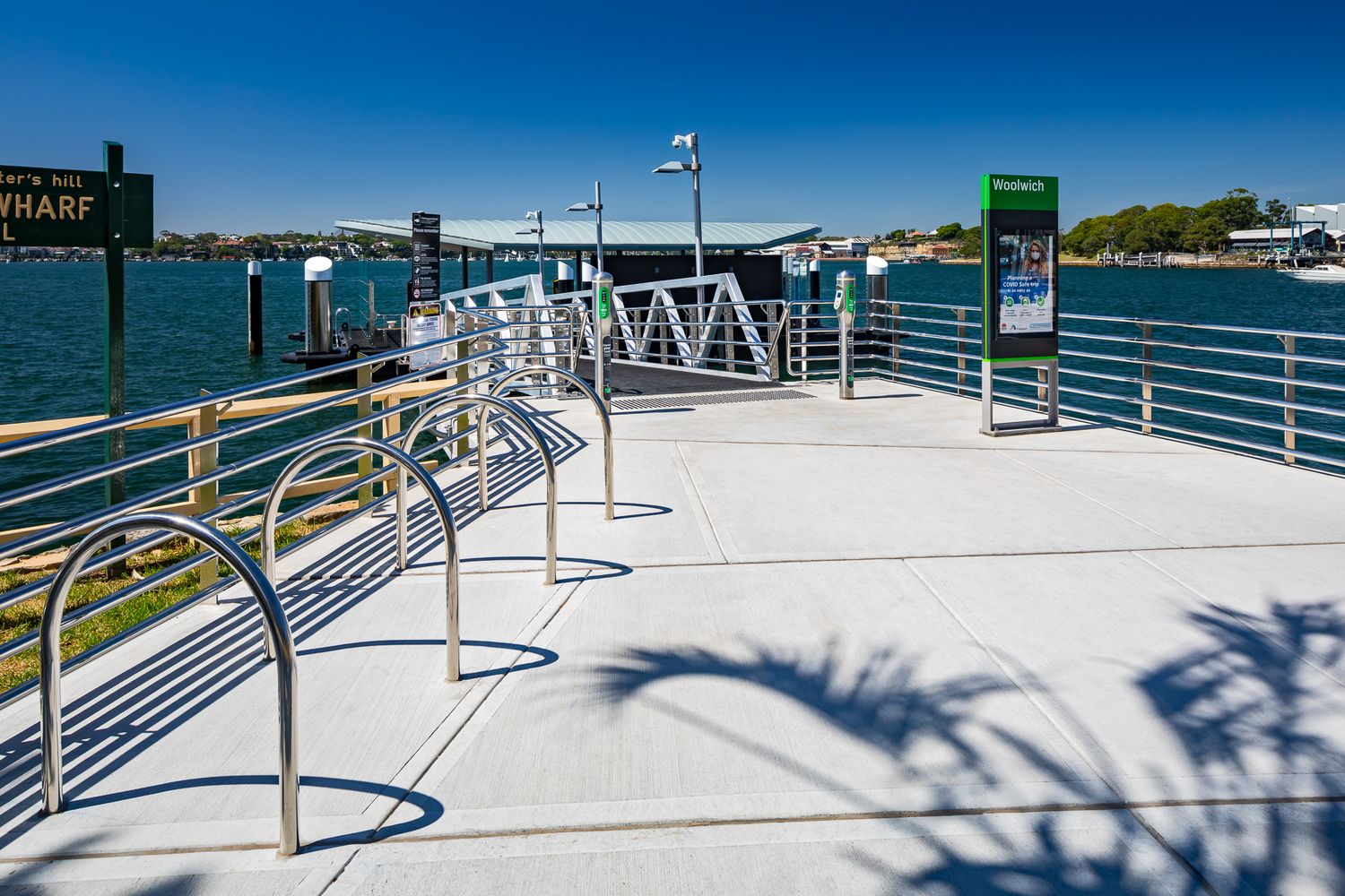 Sunny ferry wharf deck with bicycle racks, a timetable sign and a gangway leading to the pontoon over blue harbour water.