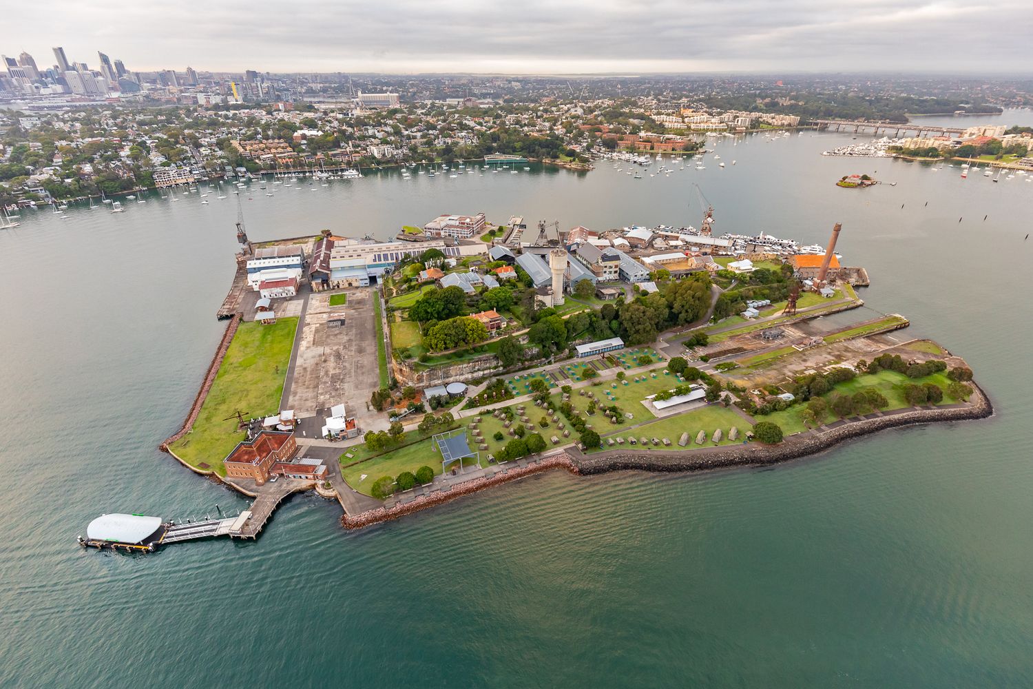 Aerial view of a green island with historic buildings and a small wharf surrounded by harbour waters and a distant city skyline.