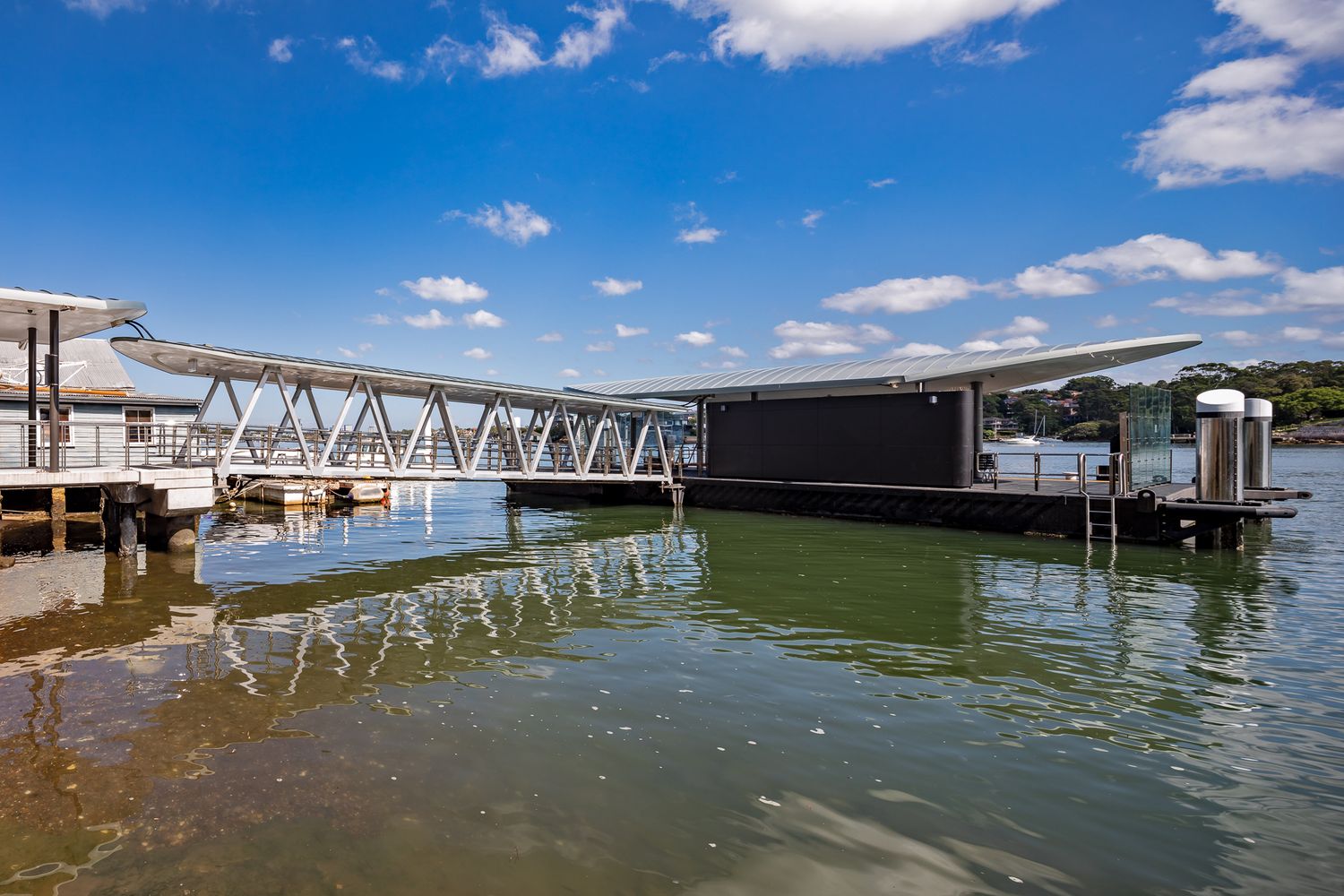 Ferry wharf pontoon and steel truss gangway reflected in still green harbour water beneath a blue sky with scattered clouds.