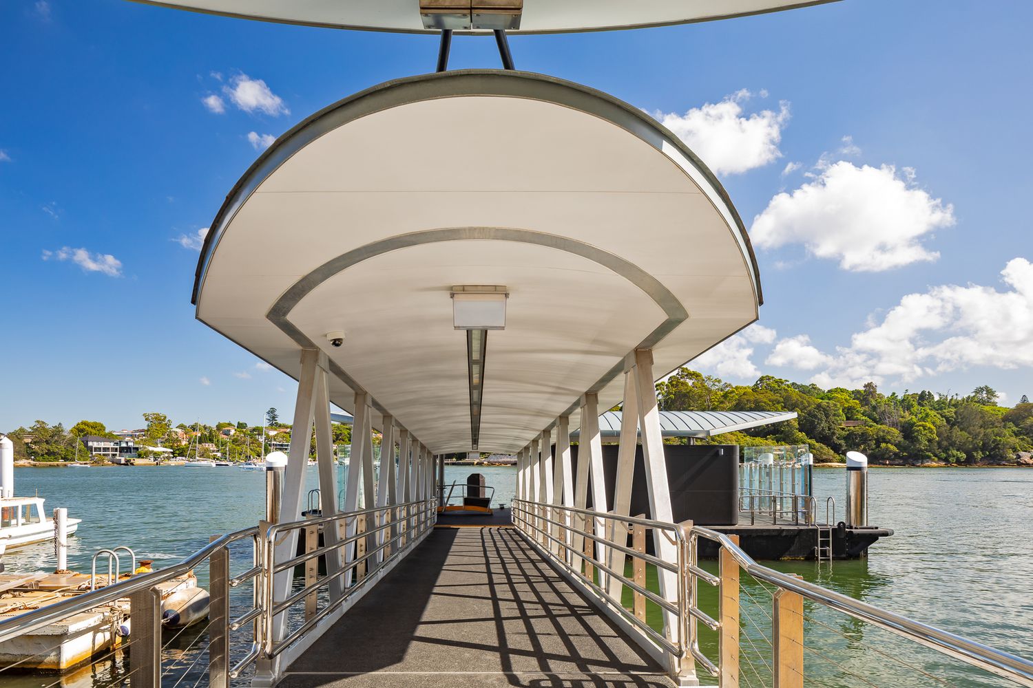 Covered ferry wharf walkway with curved white canopy and railings leading out over sparkling harbour water under a blue sky.