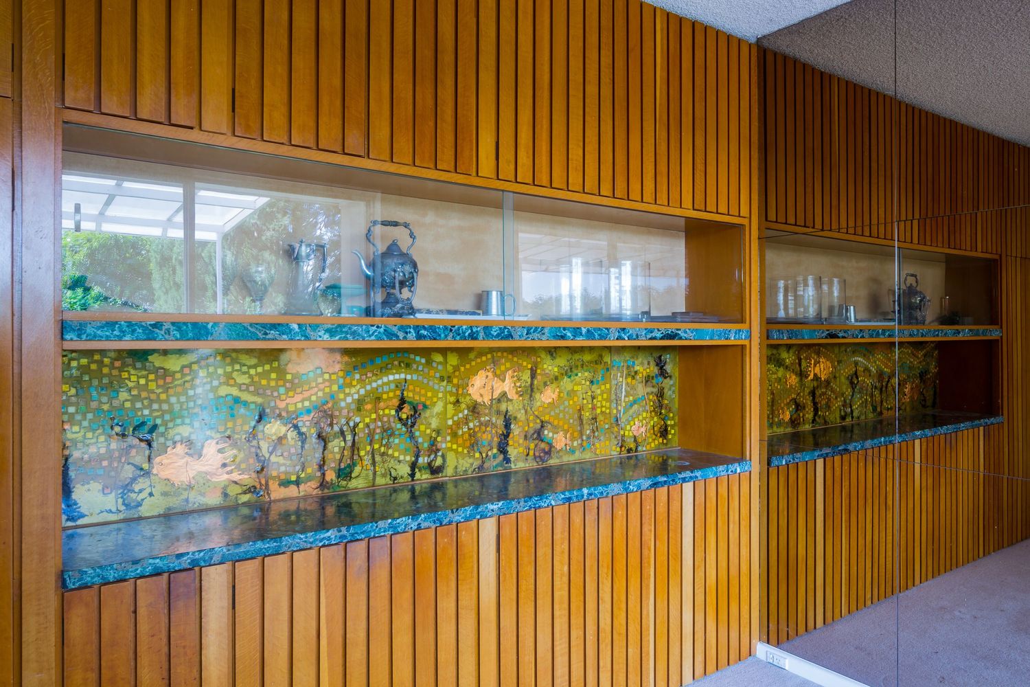 Built-in timber-panelled display cabinet with illuminated glass shelves showcasing colourful mosaic artwork and decorative vessels.