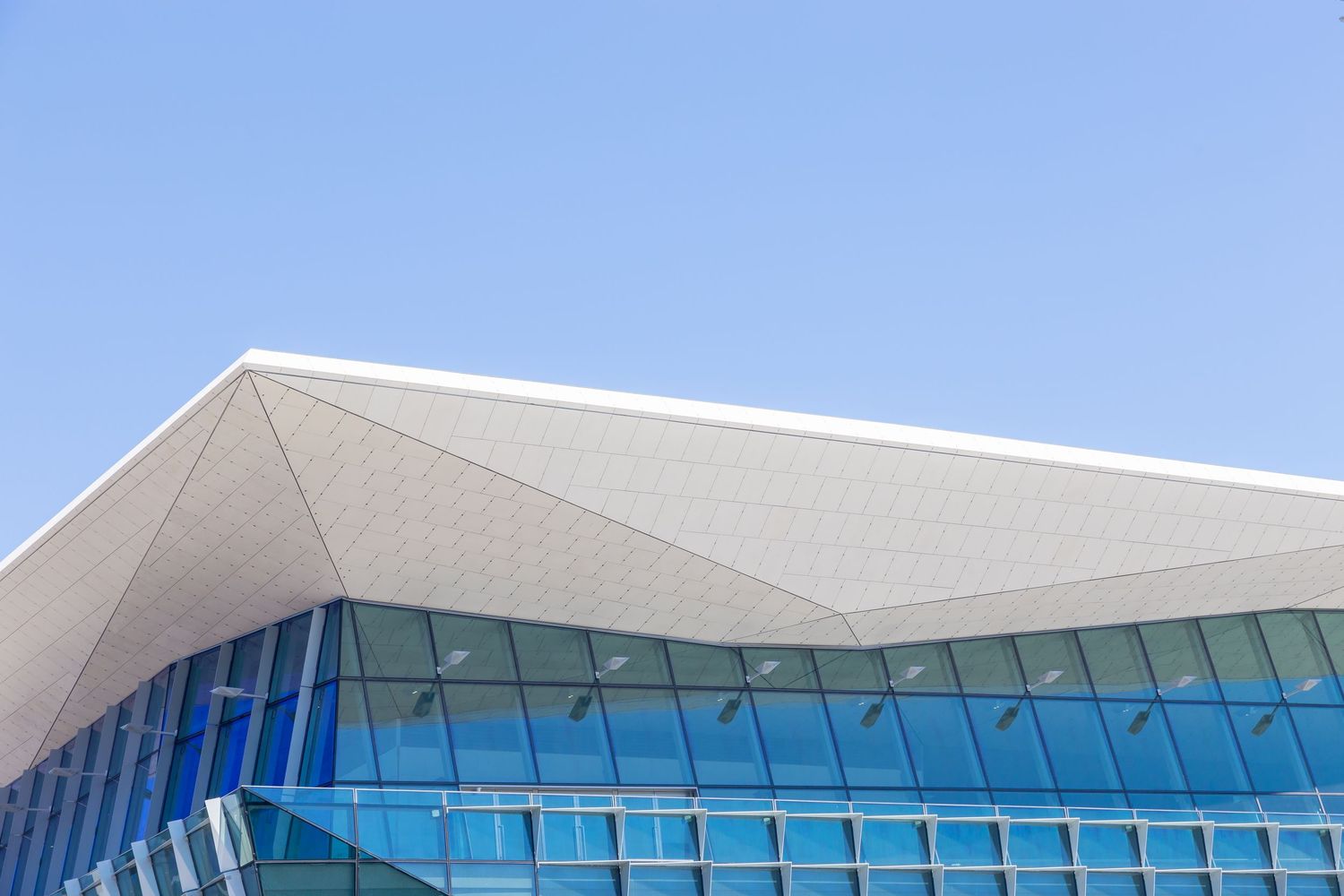 Close view of a modern building's angular white roof canopy above a blue glass curtain wall under a clear blue sky.