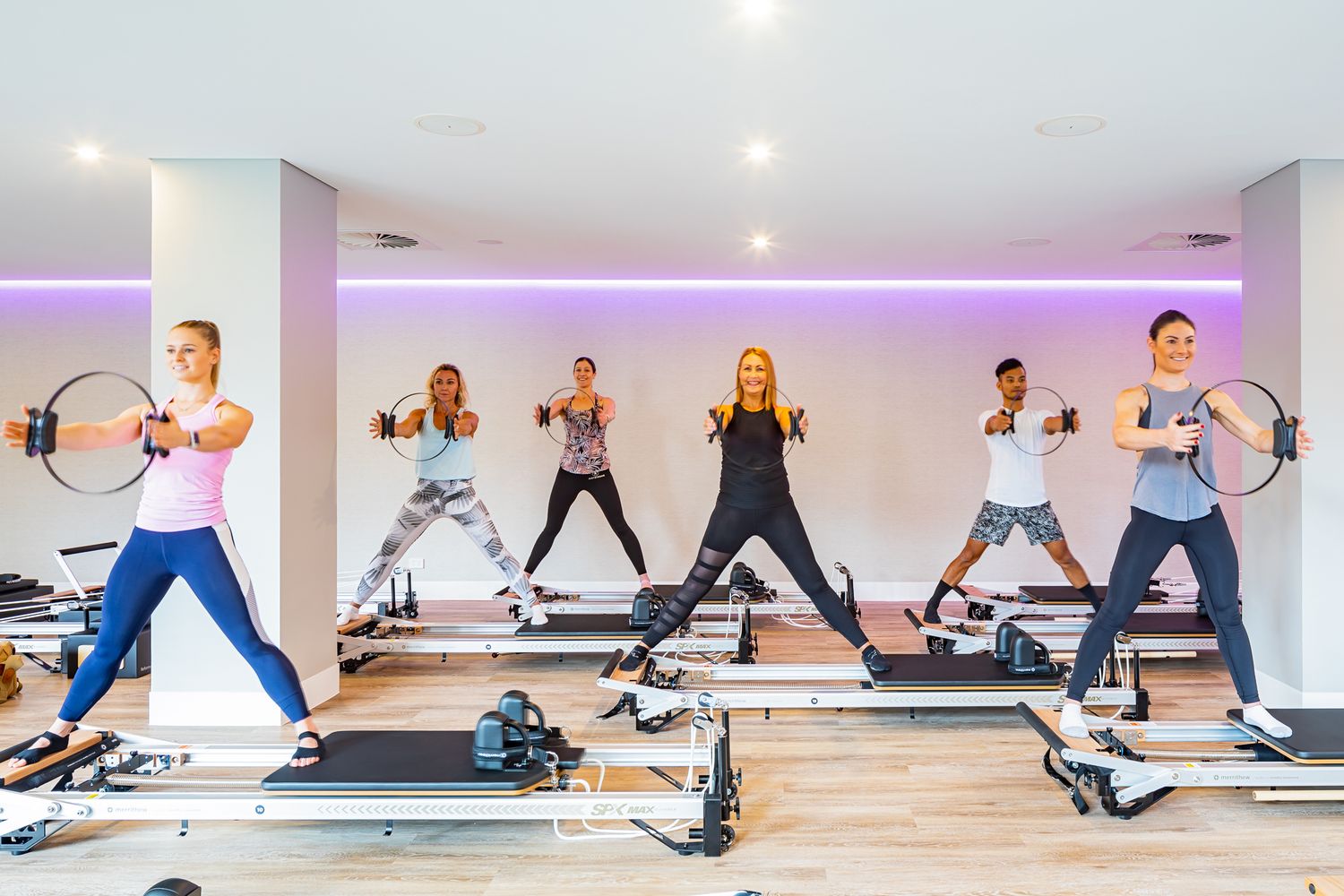 Six women exercise in a bright studio on reformer Pilates machines, each holding a Pilates ring during a group fitness class.