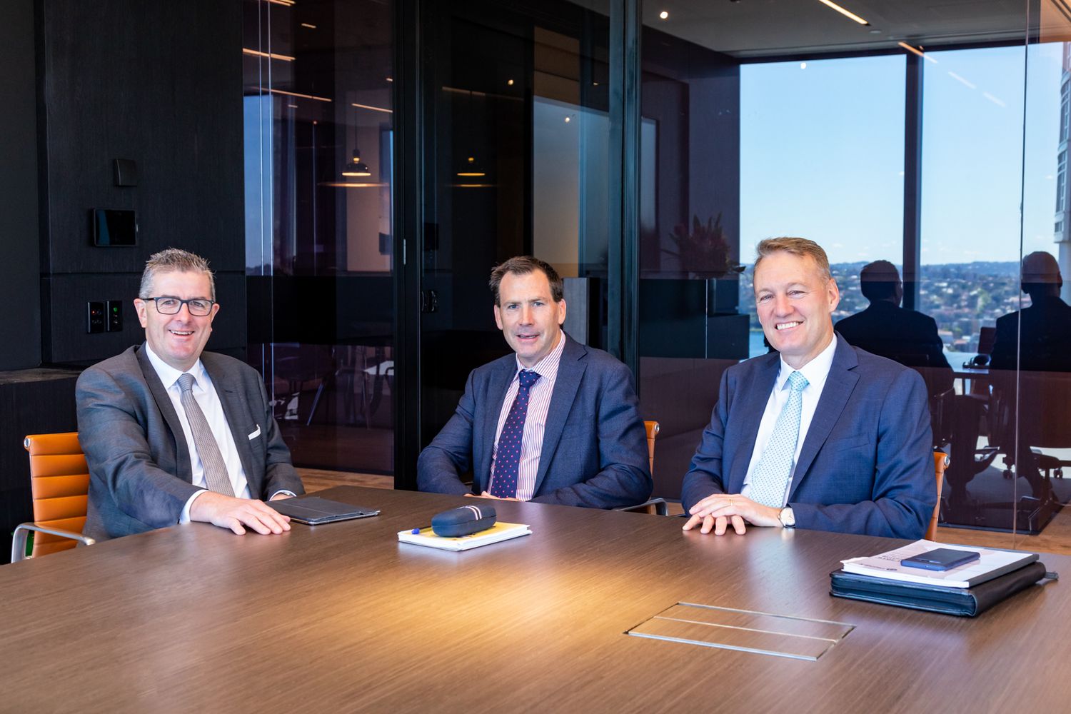 Three men in business suits sit smiling at a wooden boardroom table in a modern glass-walled office with city views.